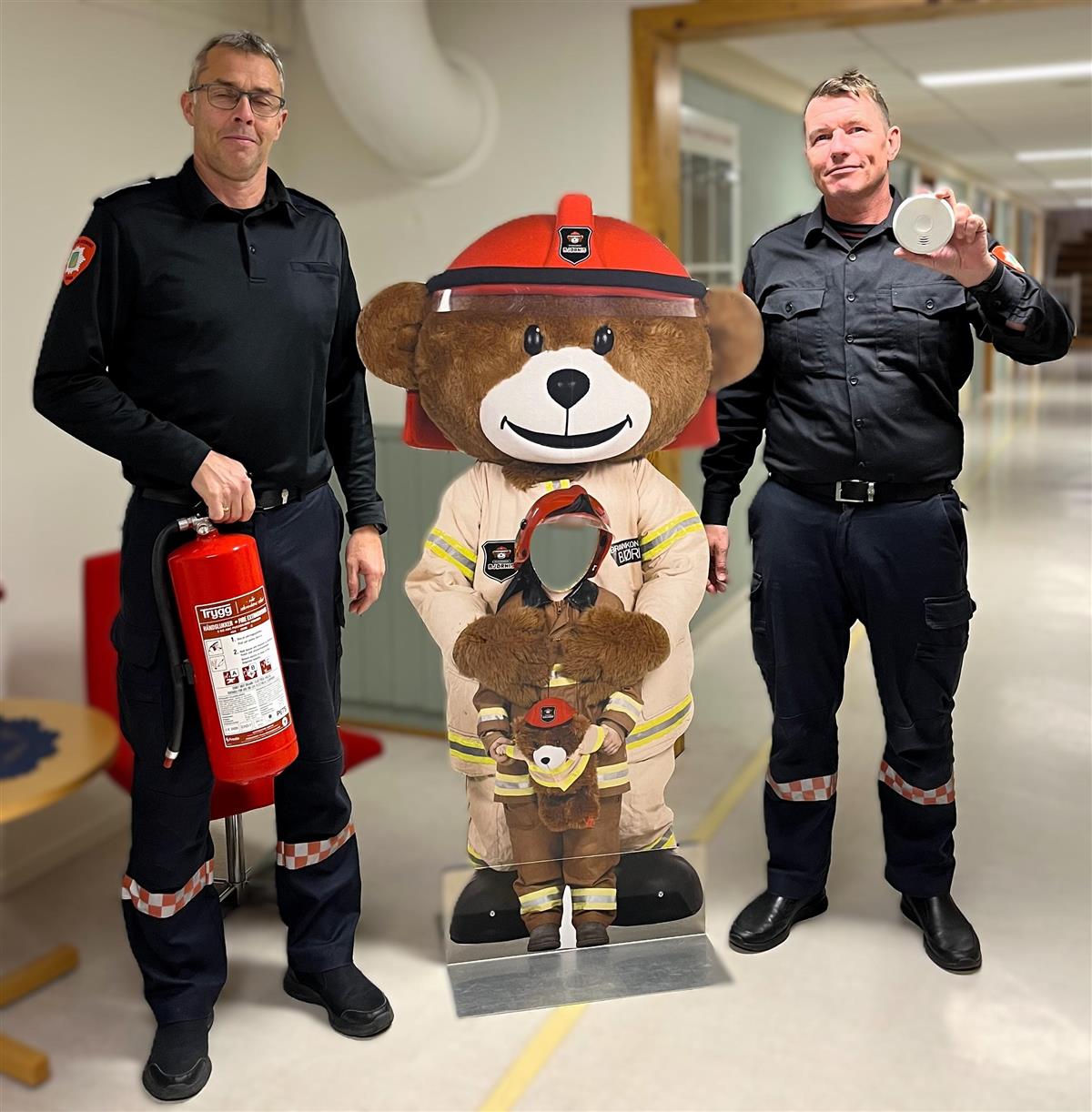 To brannmenn med en stor bamse mellom seg. Den ene brannmannen holder et slokkeapparat, den andre holder en røyvarsler.  - Klikk for stort bilde