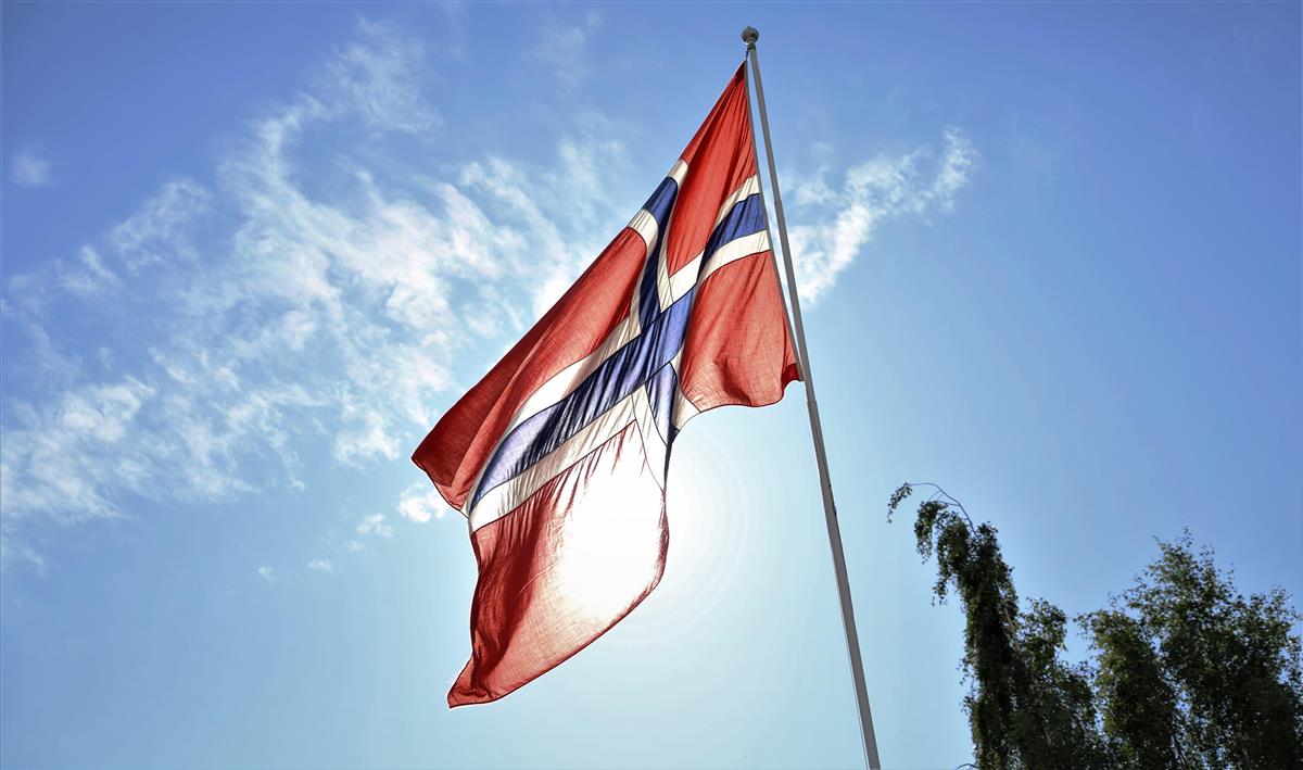Norges flagg som henger i toppen av en flaggstang, med blå himmel og solen bak flagget. Foto - Klikk for stort bilde